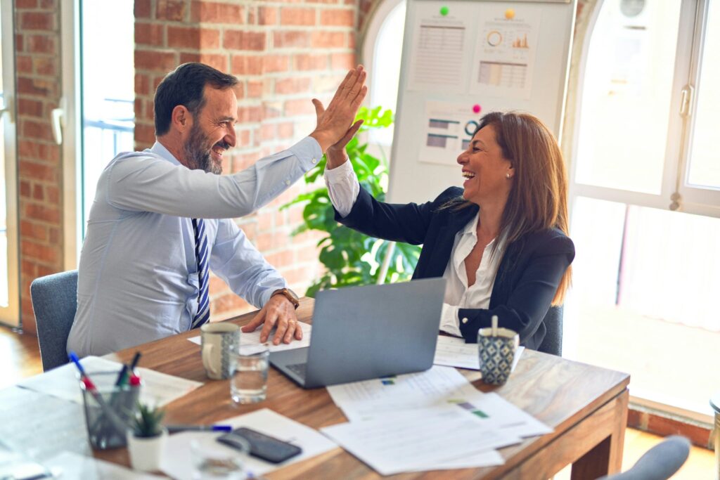Homme en chemise blanche et femme en chandail noir assis dans un bureau lumineux, se donnant un high‑five avec un sourire, illustrant une collaboration professionnelle et un soutien aux entreprises.